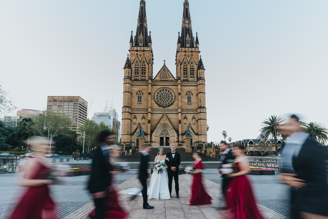 Couple standing still outside St Mary's Cathedral as the bridal party rushes past