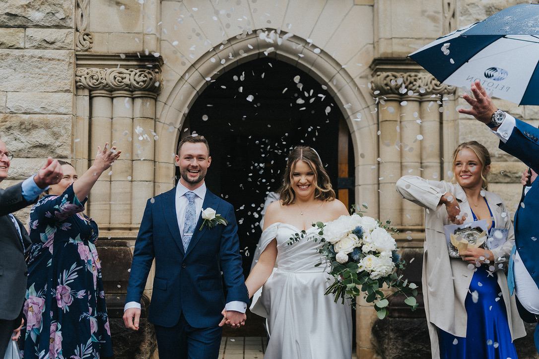 Newlyweds walking out of a sandstone church through falling petals