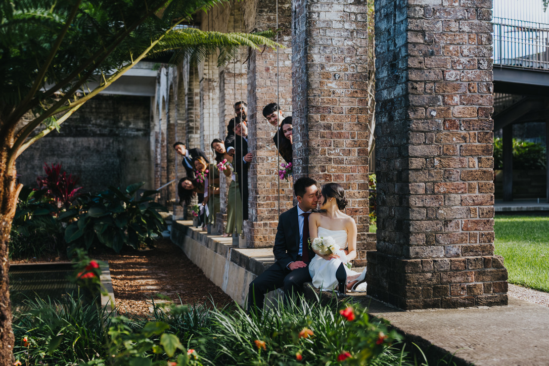 Bridal party peeking around brick pillars at the couple